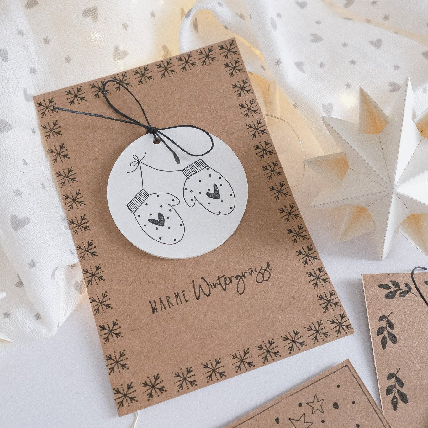 Anwendungsbeispiel für Stempel mit Handschuhen als Motiv. Karte in Kraftpapier mit Schneeflocken als Rahmenmotiv. Darauf ein gegossener Anhänger aus Gips bestempelt mit den Handschuhen. Text Warme Wintergrüsse.