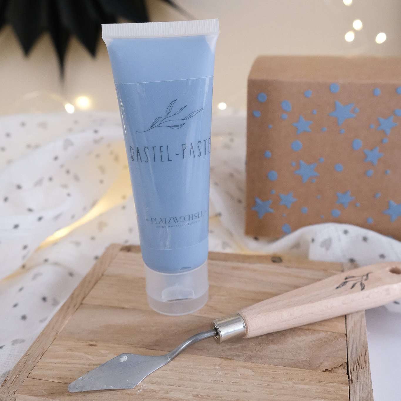 Produktfoto von einer Tube mit Strukturpaste in Grau Blau, einem Künstlerspachtel und Bastelbeispiel einer Geschenkbox verziert mit der Strukturpaste.